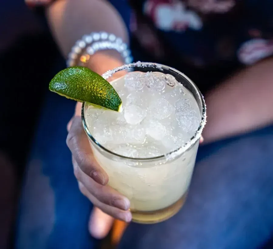 Hand holding a clear glass margarita with a salted rim and lime.