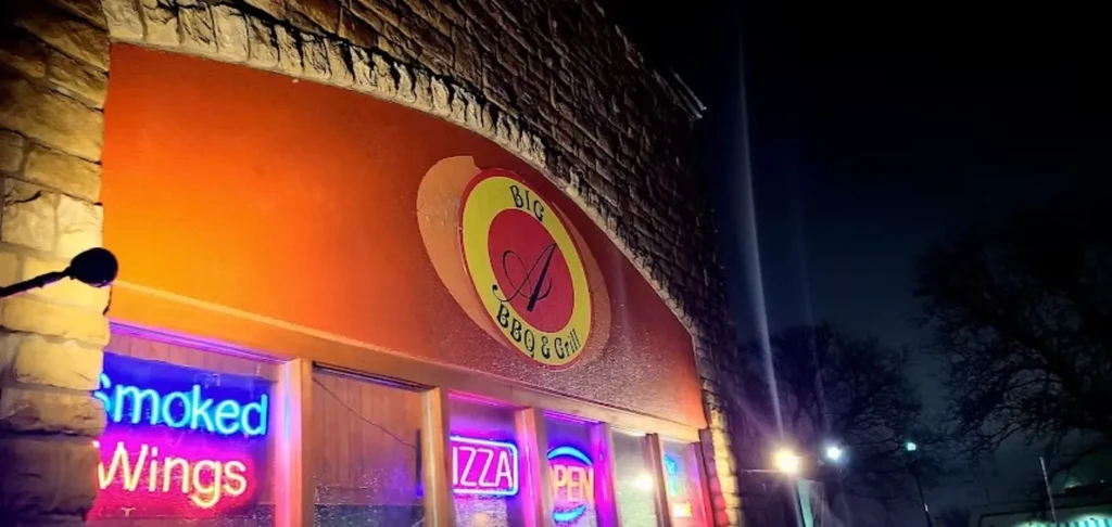 Restaurant facade at night: stone, orange wall, logo, and neon signs.