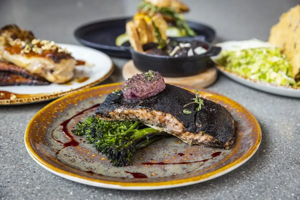 Foreground: seared salmon with broccoli and topping. Multiple other plated dishes.