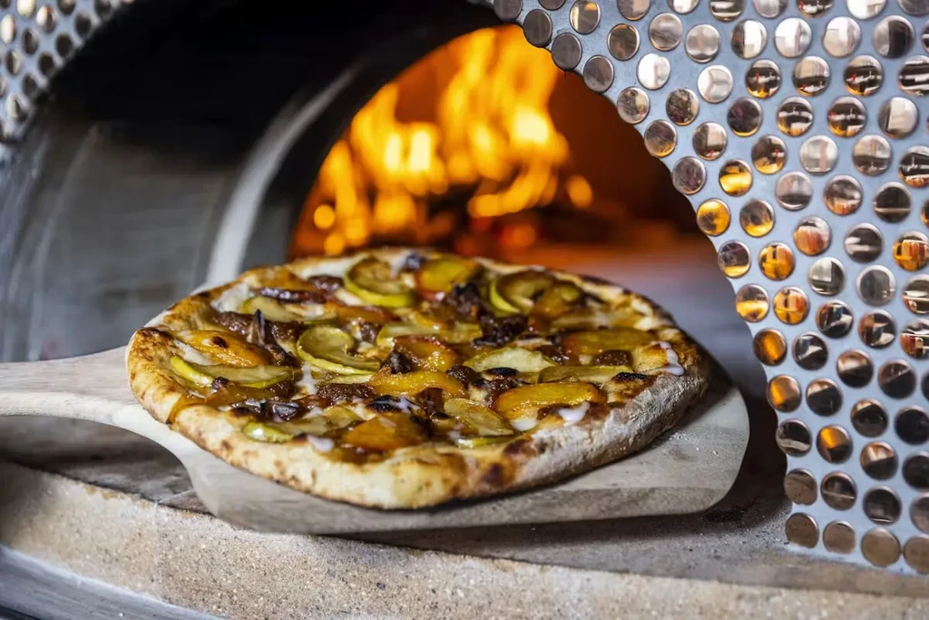 Freshly baked fruit pizza on a peel, near wood-fired oven flames.