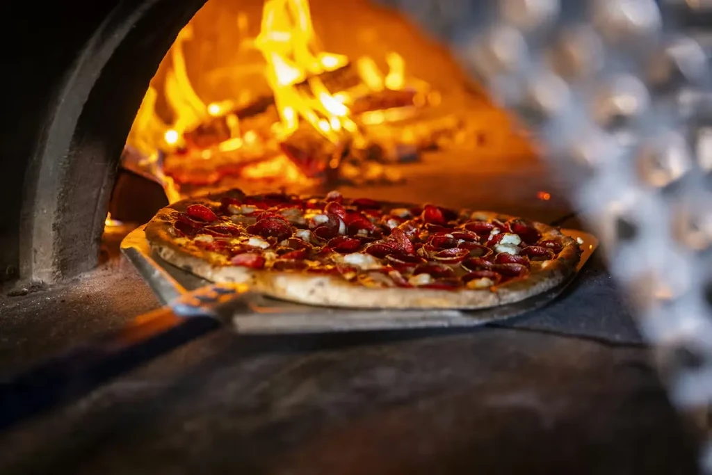 Pepperoni pizza cooking in a rustic, blazing wood-fired oven.