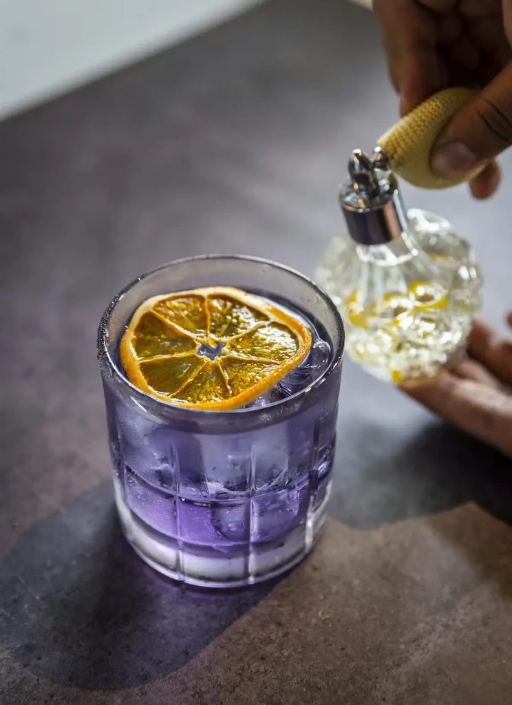 Hand mists purple cocktail with orange slice using vintage atomizer.
