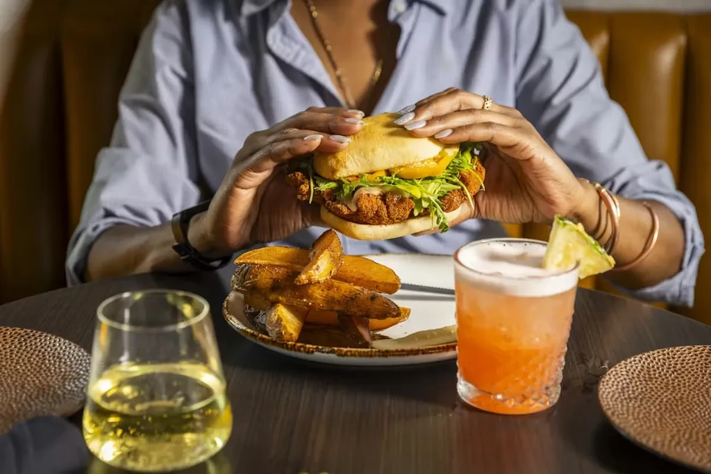 Hands hold a golden-fried sandwich, potato wedges, orange cocktail, and wine.