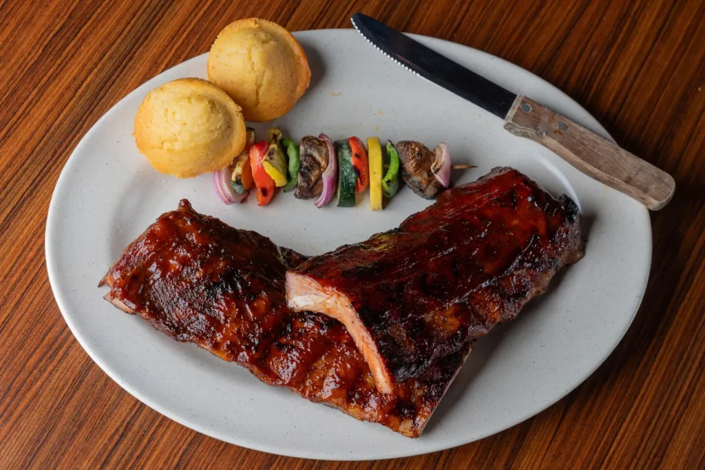 Plate of saucy BBQ ribs, cornbread, grilled vegetables, and knife.