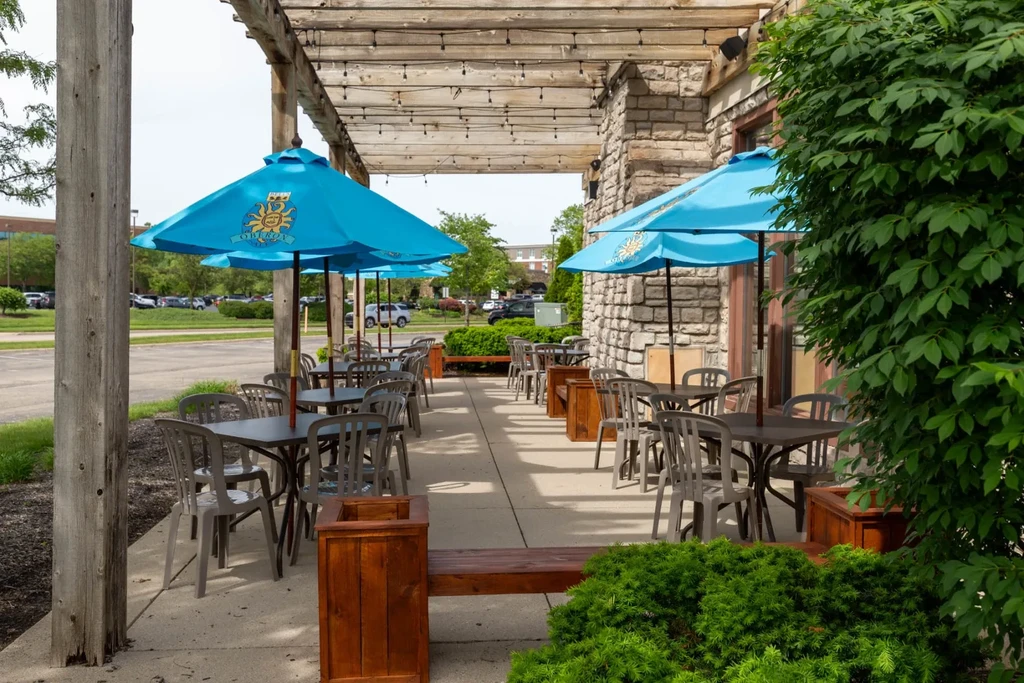 Sunny outdoor restaurant patio with blue umbrellas under a wooden pergola.