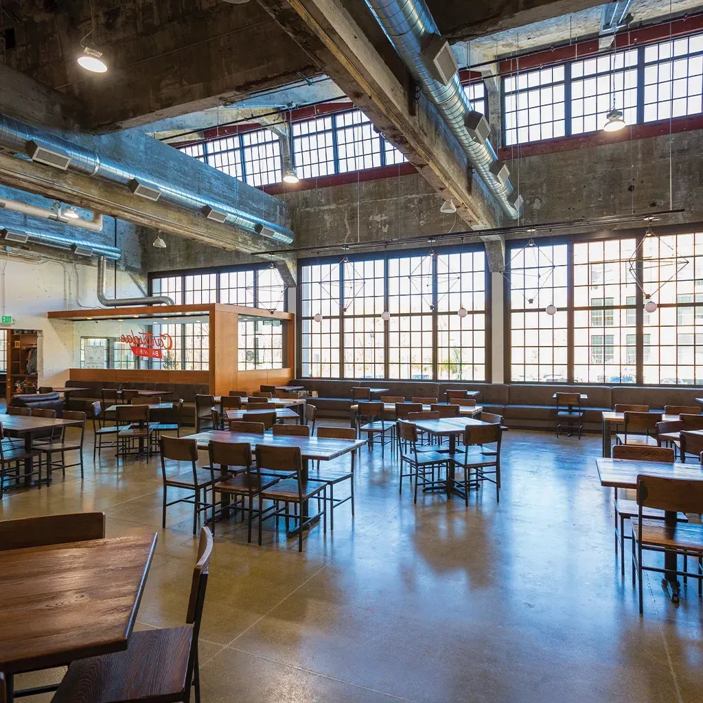 Spacious, empty industrial-chic restaurant with concrete, tall windows, and wooden furniture.