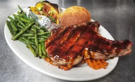 Plated glazed pork chop, loaded potato, green beans, sweet potato fries.