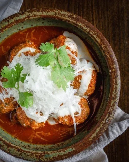 Rustic bowl of meatballs with red and white sauces, cheese, cilantro.