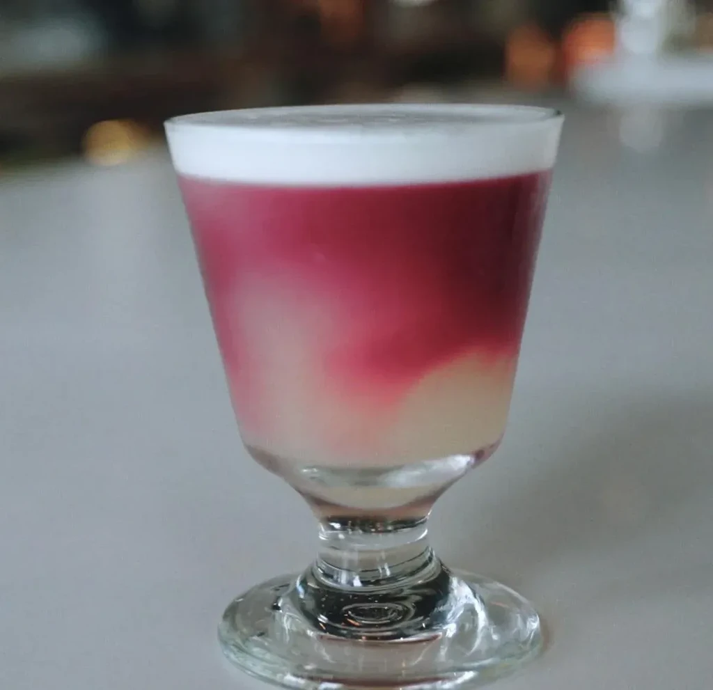 Layered pink drink in stemmed glass with white foam.