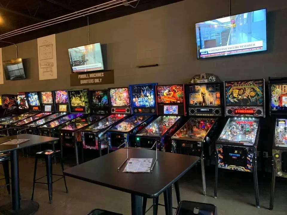 A dim arcade bar with illuminated pinball machines and news displays.
