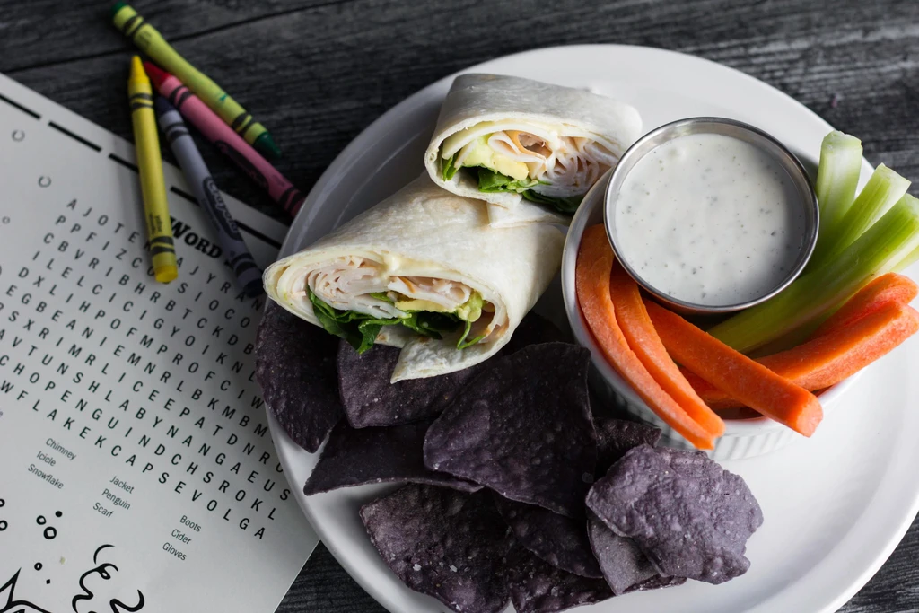Overhead: Turkey wrap, chips, vegetables, dip, and crayons on a table.