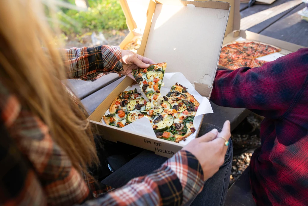 Hands reaching for a small vegetable pizza in a cardboard box outdoors.