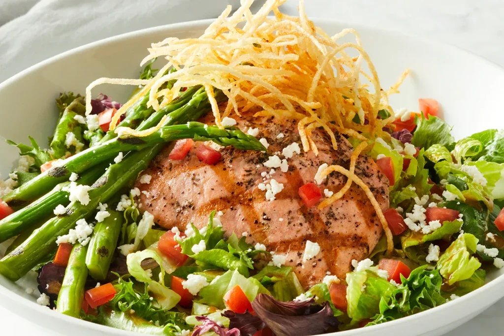 Grilled salmon salad with asparagus, tomatoes, cheese, and crispy strips in a bowl.