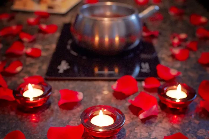 Lit votive candles, red rose petals, and fondue pot on a table.