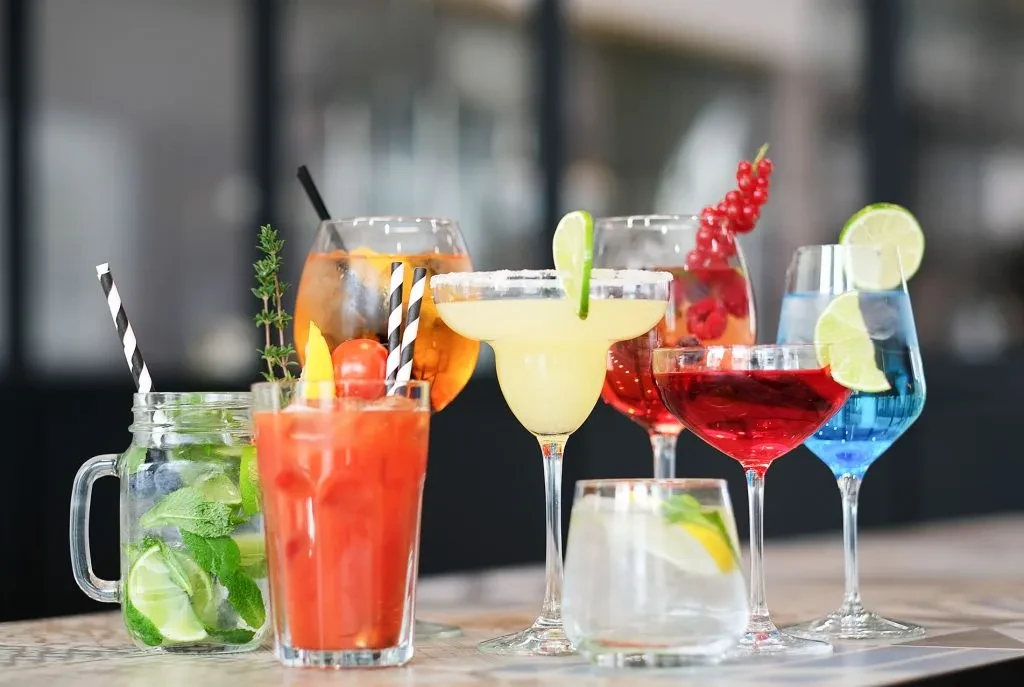 Eight colorful cocktails garnished with various fruits and herbs on a counter.