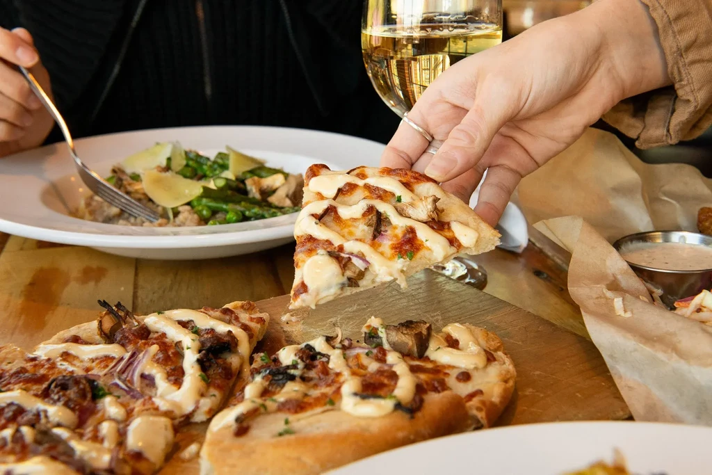 Hands sharing pizza, asparagus, wine, and sides at a wooden table.