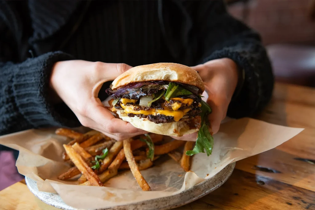 Hands holding a bitten double cheeseburger with fries on a rustic plate.