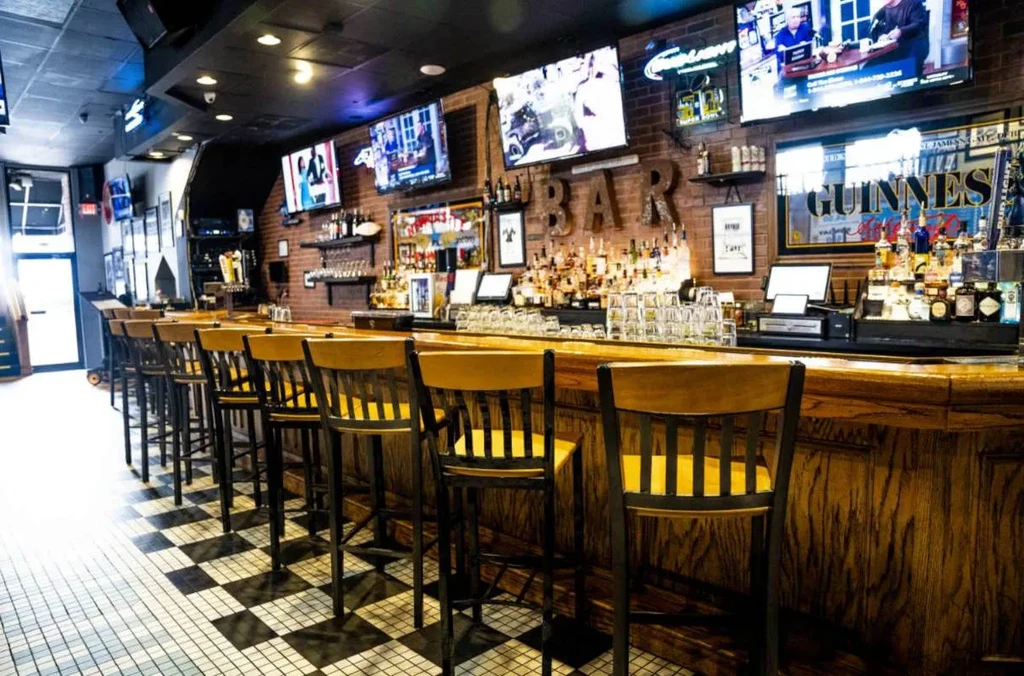 Modern bar interior featuring a wooden counter, stools, and several TVs.