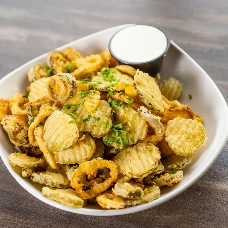 Close-up of fried pickles and onion rings with herbs and dipping sauce.