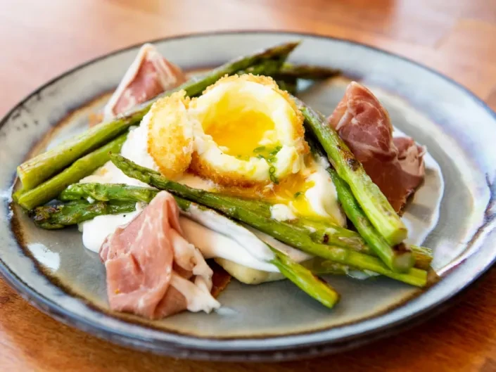 Crispy fried egg, runny yolk, asparagus, and prosciutto on a rustic plate.