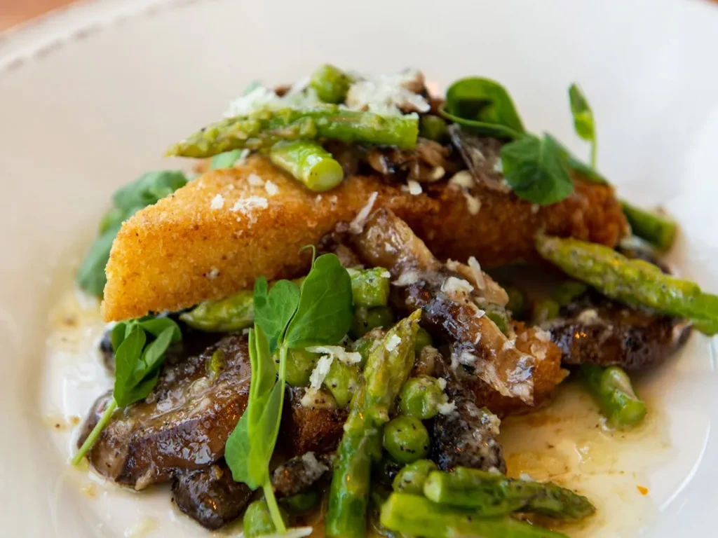 Golden-brown fried cutlet on mushrooms, asparagus, and peas with green garnish.