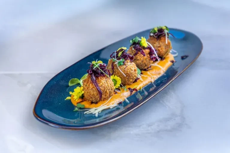 Four golden croquettes on blue dish with orange sauce, microgreens, flowers.