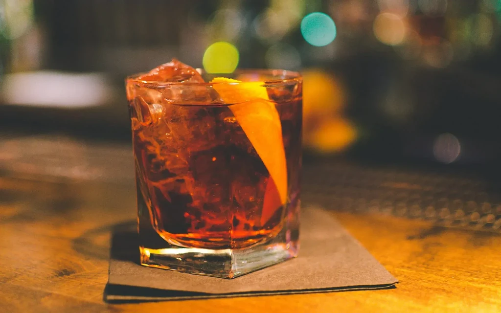 Reddish-brown cocktail with ice and orange peel in a dim bar.