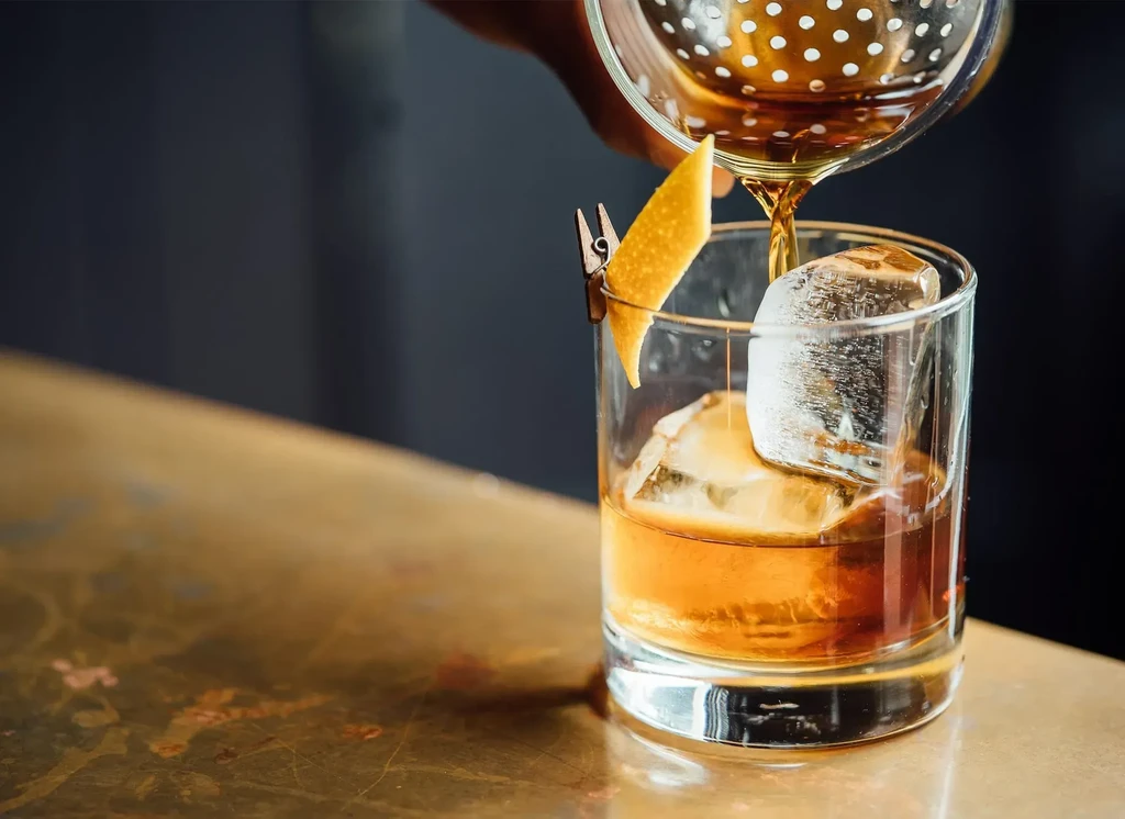Amber cocktail poured into glass with ice, garnished with citrus peel.