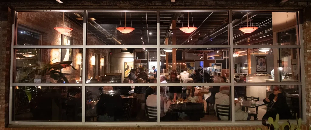 Bustling restaurant interior with warm lighting, seen through glass at night.