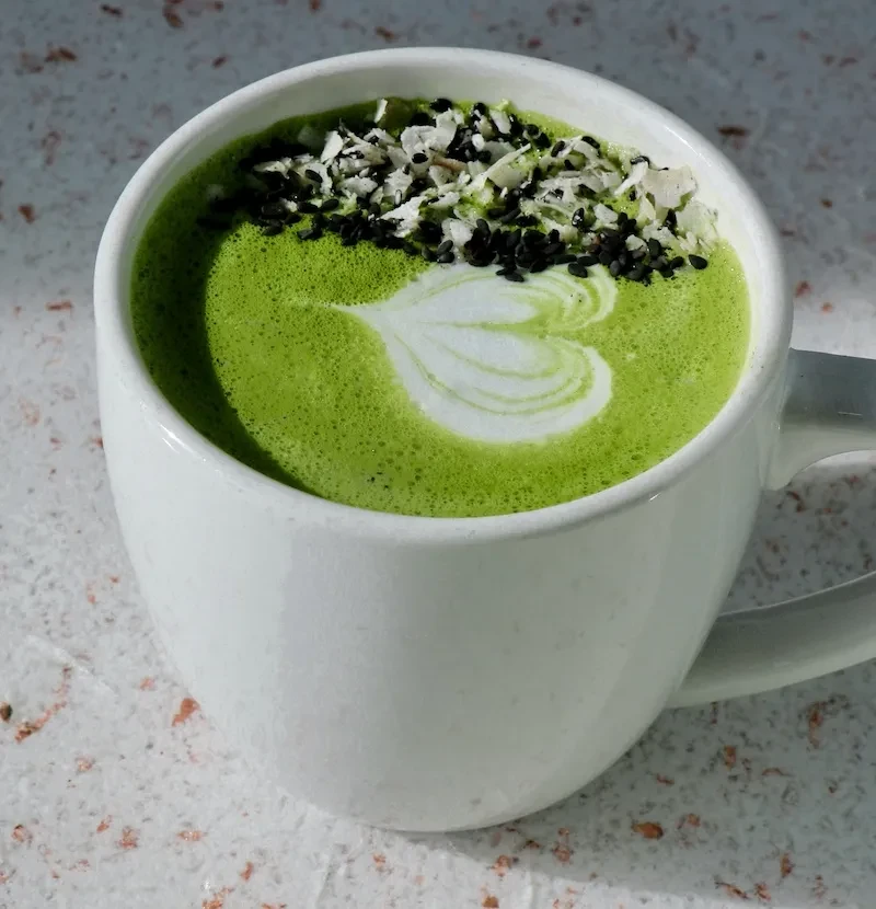 Green matcha latte with heart latte art, sesame, and coconut flakes.