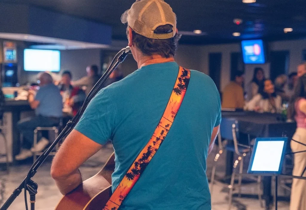 Person from behind plays acoustic guitar in a dimly lit bar.