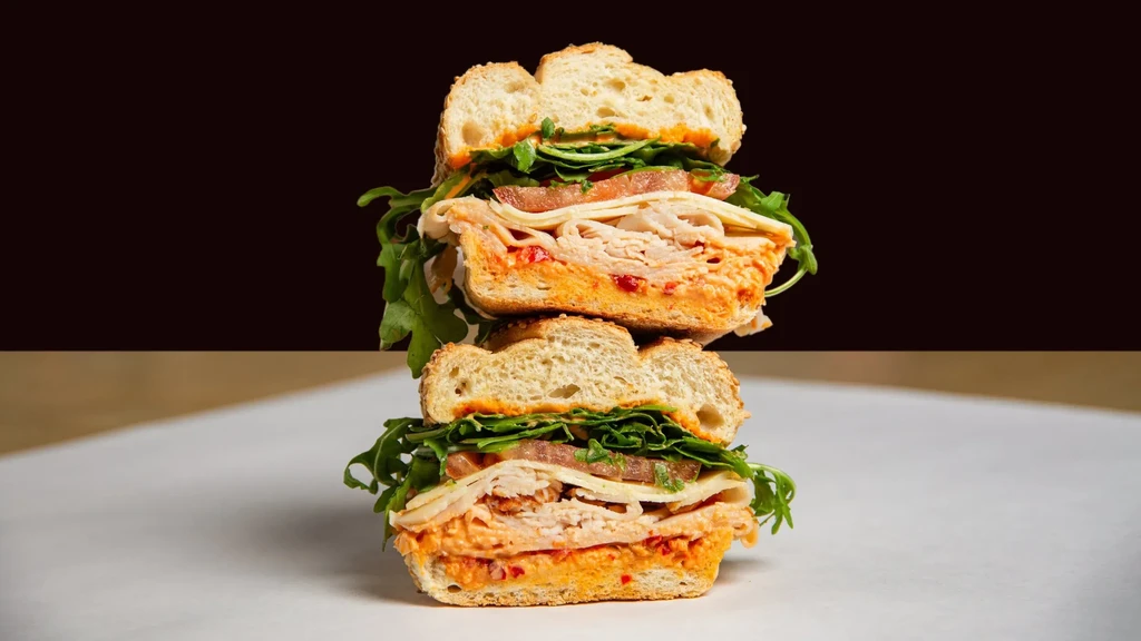 Cut, stacked turkey sandwich showing layers of cheese, arugula, tomato, spread.