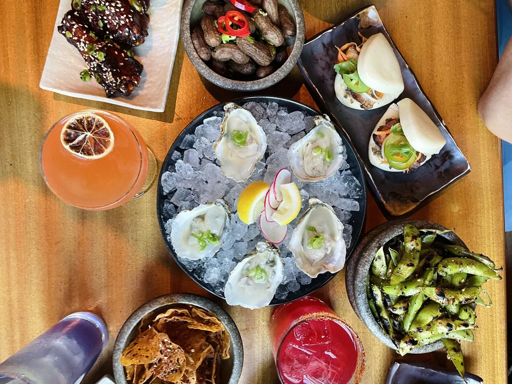 Overhead view of a table with oysters, bao buns, wings, and cocktails.