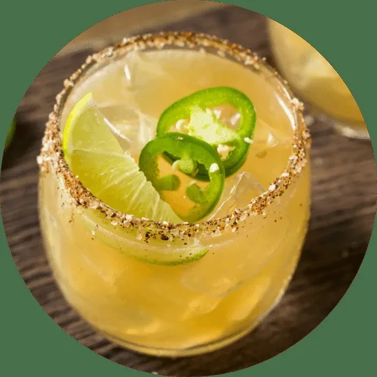 Pale yellow cocktail with ice, chili-salt rim, lime wedge, and jalapeños.