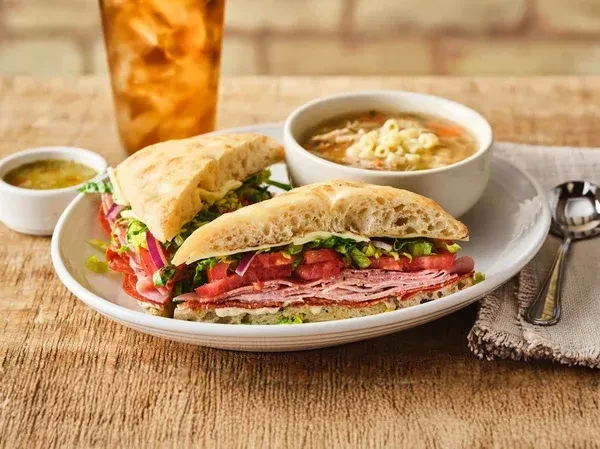 Deli sandwich, chicken noodle soup, and iced tea on a wooden table.