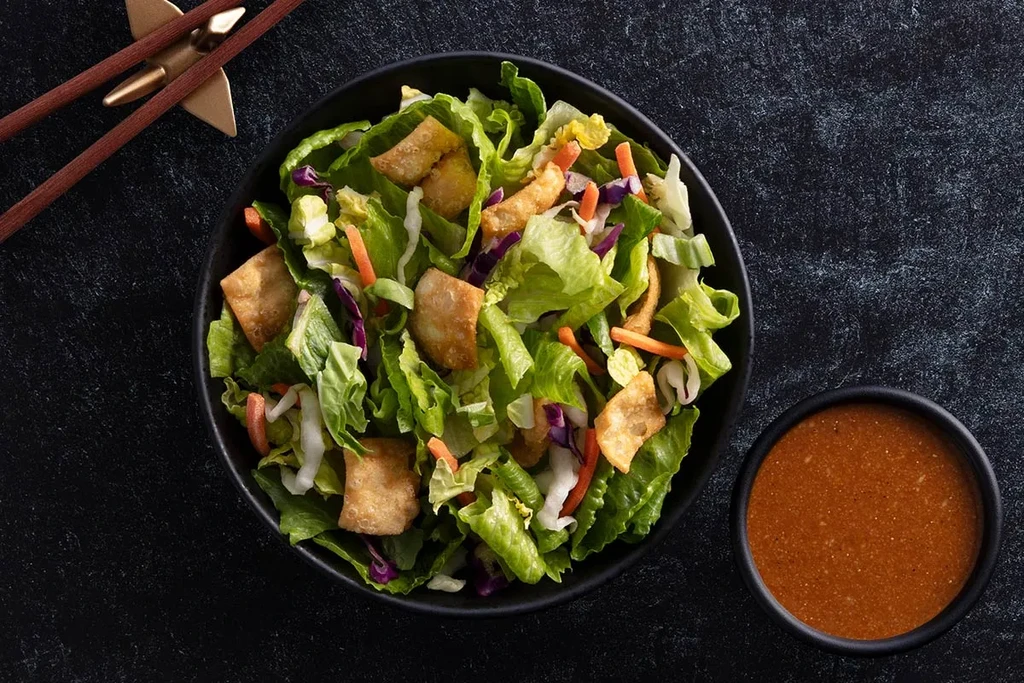 Overhead colorful salad with crispy strips, dressing, chopsticks on dark surface.