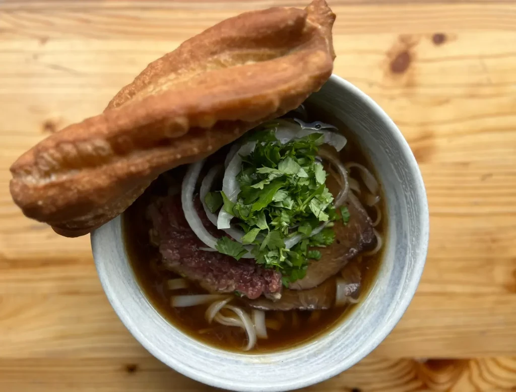Top-down pho bowl with meat, noodles, herbs, and fried dough.