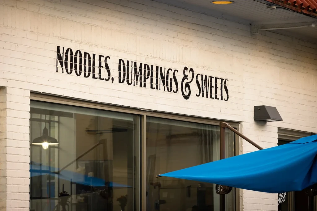 Restaurant storefront with "NOODLES, DUMPLINGS & SWEETS" sign and blue umbrella.