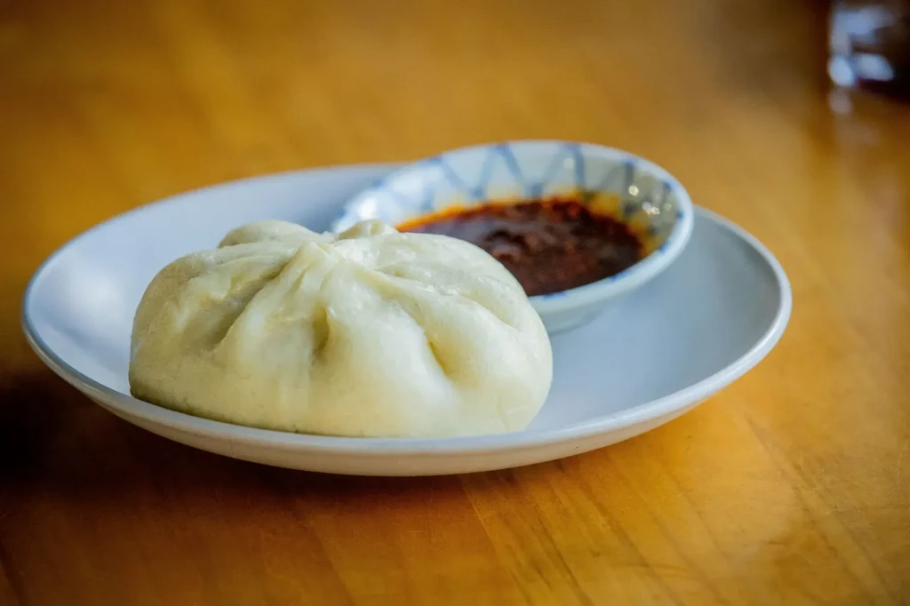 Light steamed bun with pleats, on a white plate with chili sauce.