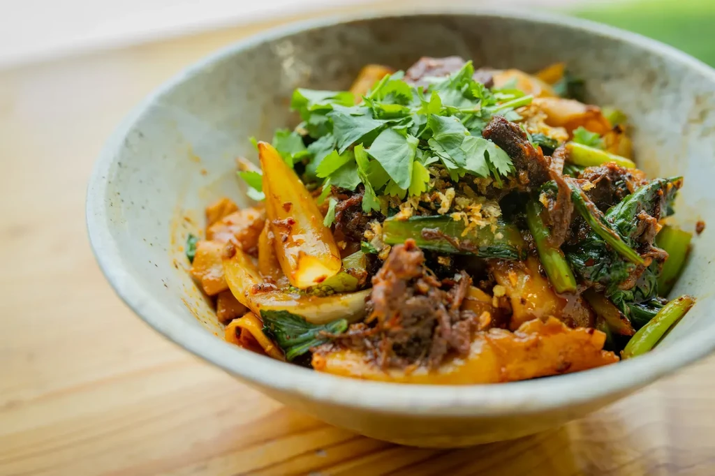 Bowl of stir-fried noodles with meat, vegetables, cilantro, and sauce.
