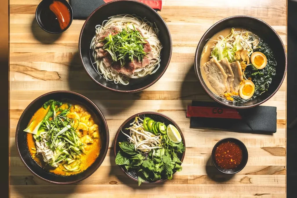 Overhead flat lay of four Asian noodle and soup bowls with condiments.