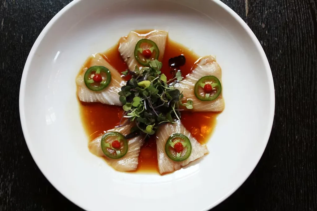 Plated raw fish crudo with jalapeño, chili, microgreens, and sauce.