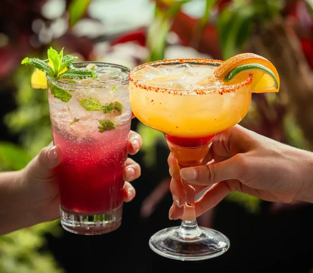 Two hands hold a pink fizzy cocktail and an orange margarita.