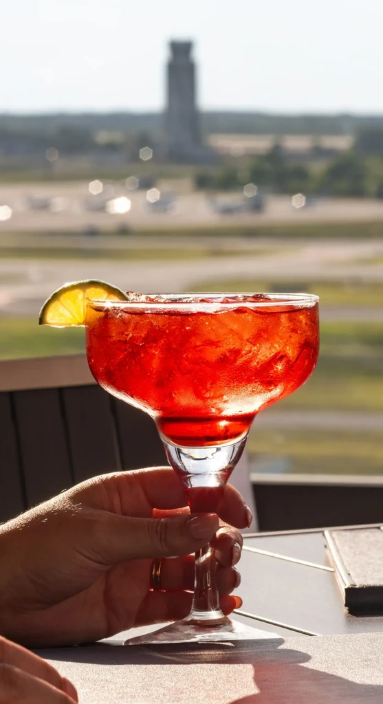 Hand holds red cocktail with lime outdoors, blurred tower and airfield.