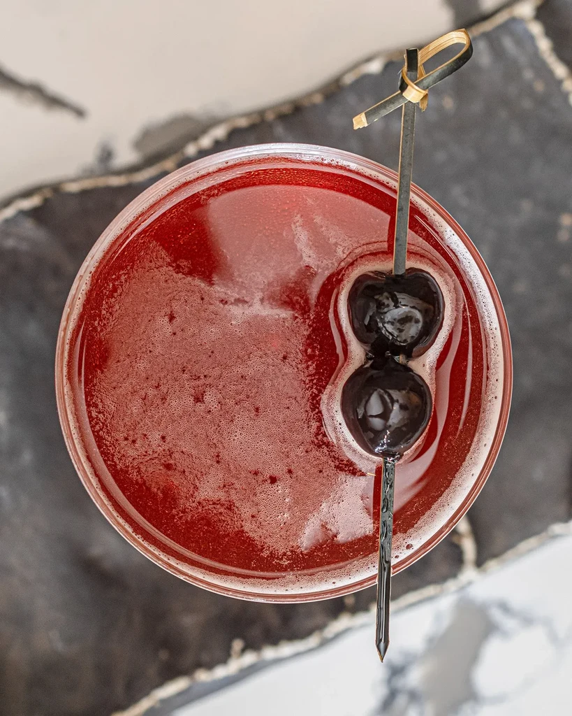 Overhead view of a red cocktail with two dark cherries on a skewer.