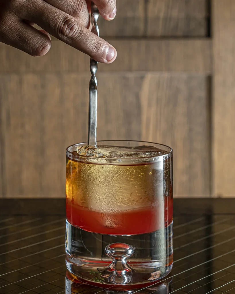 Hand stirring a colorful layered drink with a large ice sphere.