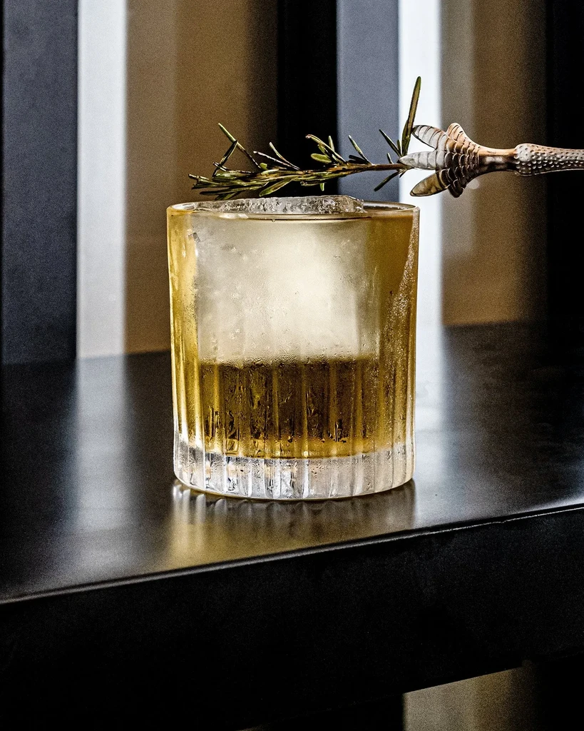 A cocktail in a glass with a rosemary garnish and claw-shaped stirrer.