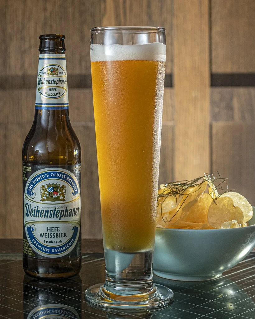 A bottle of Weihenstephaner beer next to a glass of beer and chips.