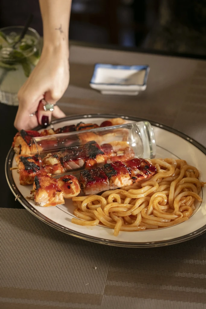 A person's hand placing a glass bottle over salmon skewers and noodles.