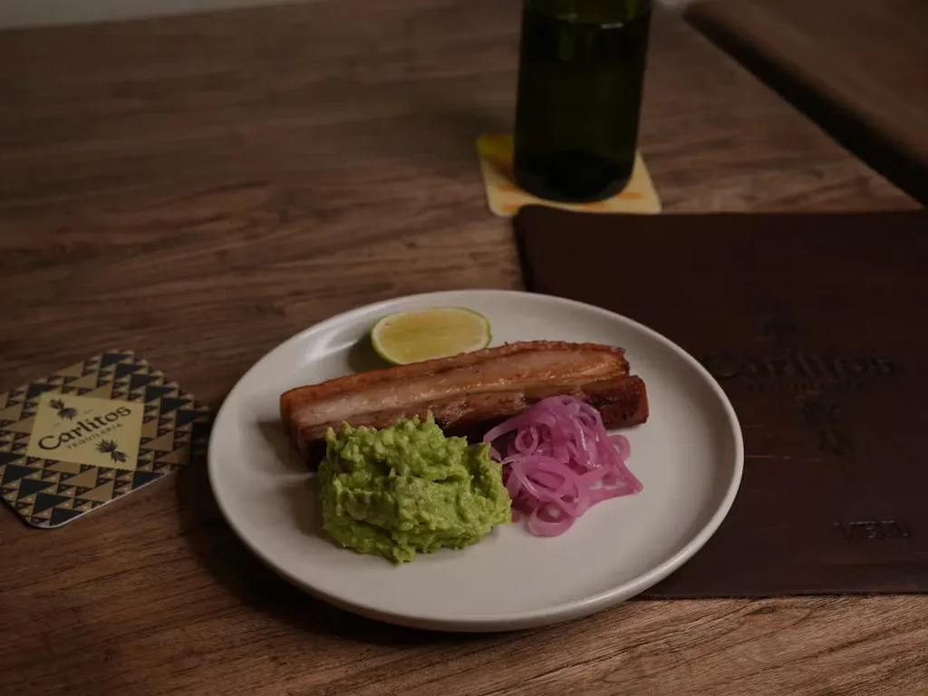 Pork belly with guacamole, pickled onions, and lime on a plate.